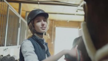 İki genç kadın birlikte at tımarlıyor, ahırda çalışırken gülümsüyor ve sohbet ediyorlar.