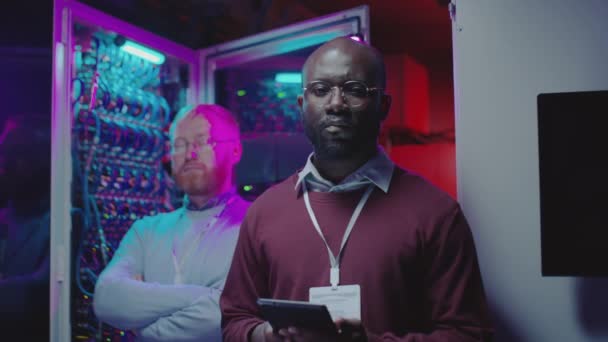 アフリカ系アメリカ人と白人のデータセンター技術者の肖像ネオンと暗いサーバールームでカメラのポーズ — ストック動画