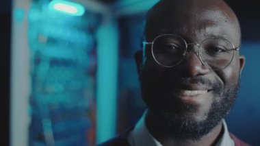 Afro-Amerikan veri merkezi mühendisinin portresi, karanlık sunucu odasında mavi ışıkla poz verirken kameraya gülümsüyor.