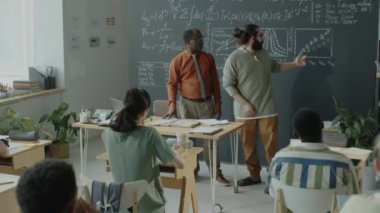 Genç melez öğrenci yazı tahtasının yanında duruyor ve ders sırasında akranlarının ve Afro-Amerikan profesörün önünde konuşma yapıyor.