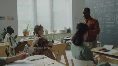 Orta yaşlı Afro-Amerikan profesör sınıfta sıraların arasında yürüyor ve lisede ders verirken çeşitli öğrencilerle konuşuyor.