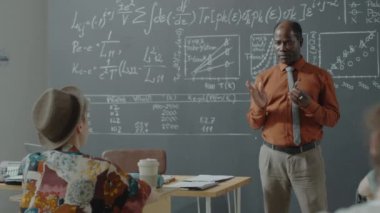 Afro-Amerikan profesör tahtadaki grafiklere ve formüllere işaret ediyor ve üniversitede matematik dersi verirken öğrencilere hitap ediyor.
