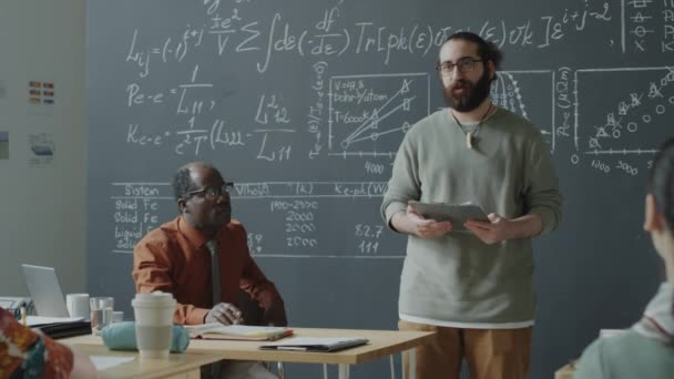 在大学课堂上 有胡子的学生站在黑板旁边 一边在剪贴板上看书 一边向集体教授和非洲裔美国人教授演讲 — 图库视频影像