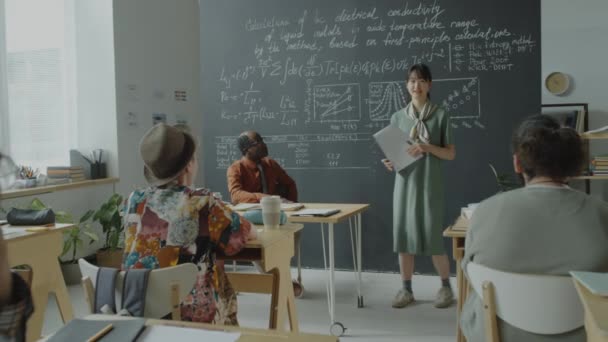 アジアの女の子は 大学で数学の授業中に様々なグループの仲間とアフリカ系アメリカ人の教授に論文を保持し 数式やグラフを説明 — ストック動画