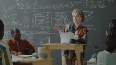 Protez kollu genç kız öğrenci kürsüde duruyor, gazete okuyor ve üniversitede matematik dersinde sunum yaparken grup arkadaşları ve profesörle konuşuyor.
