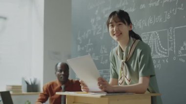 Kürsüye yaslanan, elinde kağıtlarla gülümseyen ve üniversitedeki matematik dersinde öğrencilere sunum yapan neşeli Asyalı kız.