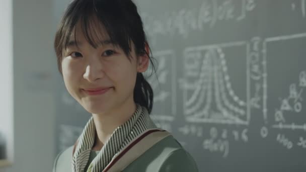 大学の教室で黒板の横にポーズをとりながらカメラを見て微笑む陽気なアジアの女の子の肖像 — ストック動画
