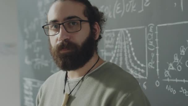 ポートレート胸アップショットのひげを生やした学生で眼鏡でカメラのために反対Chalkboardで大学の教室 — ストック動画