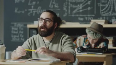 Gözlüklü sakallı erkek öğrenci, sınıfta masada oturuyor ve lisede ders sırasında öğretmeniyle konuşuyor.