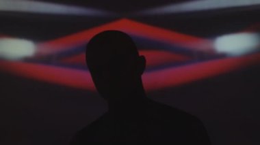 Göğsü dövmeli bir adam, baştan aşağı zincirli karanlık stüdyoda müzik eşliğinde yanıp sönen neon ışığı ve arka planda kaleydoskopik projeksiyonla dans ediyor.
