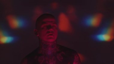 Göğsü dövmeli bir adam, baştan aşağı zincirli kameraya bakıyor ve karanlık stüdyoda kaleydoskopik projeksiyonla dans ediyor.