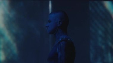 Göğsünde kulaktan kulağa zincir ve metal omuz yastığı olan bir adamın, arka planda mavi neon ışığı ve kaleydoskopik projeksiyonla karanlık stüdyoda poz vermesi.