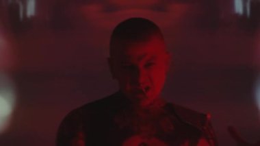 Karanlık stüdyoda neon ışığı ve kaleydoskopik projeksiyonla dans ederken kamera karşısında duygusal olarak çığlık atan, burnu kulaklarına kadar dövmeli bir adamın göğüs görüntüsü.