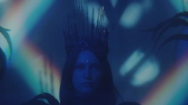 Yüzü mücevherli ve gotik tacı olan genç bir kadın. Tüylü giyinmiş. Gökkuşağı ışığı yansıtmalı karanlık bir odada duruyor.