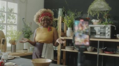 Önlüklü genç Afrikalı Amerikalı kadın mutfakta duruyor, kameraya el sallıyor ve konuşuyor.