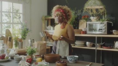 Afro-Amerikalı kadın yemek blogcusu ahşap kasede un seçiyor ve mutfakta yemek tarifi çekerken kamerada konuşuyor.