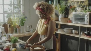 Önlüklü Afro-Amerikalı kadın ahşap kasede hamur yağlıyor ve mutfakta yemek tarifi çekerken kameraya konuşuyor.