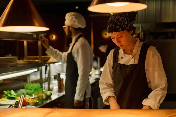 Young serious female worker of restaurant in uniform preparing food in the kitchen while standing by counter against male colleague