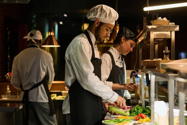 stock image Young multiethnic male chef peeling fresh zucchini while standing by workplace against female colleague mixing ingredients in pan