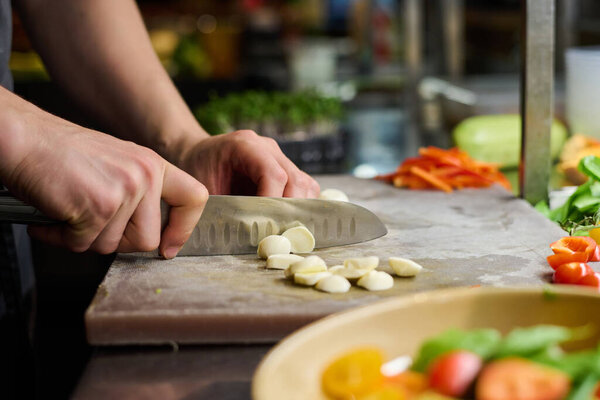 Hands of young male chef slicing pieces of fresh mozarella cheese on chopping board while preparing ingredients for salad in the kitchen