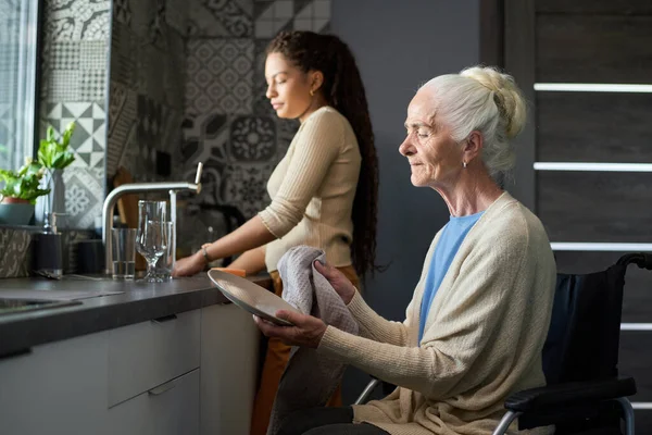 有残疾的灰发祖母坐在轮椅上与孙女一起洗碗时 用干毛巾擦拭干净盘子 — 图库照片