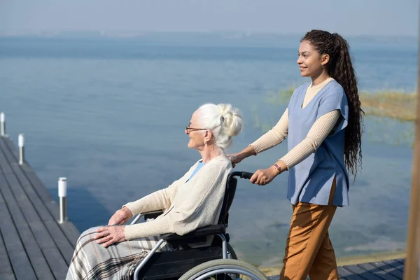 水辺で桟橋に沿って歩いている間 祖母と一緒に車椅子を押している犠牲者の若い多民族の孫娘の側面図 — ストック写真
