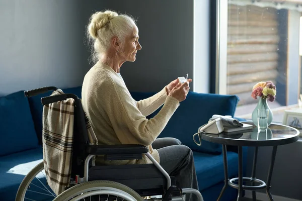 家の中で車椅子に座っている間に小さなプラスチック製の薬箱から錠剤を服用することになる障害のある退職女性の側面図 — ストック写真