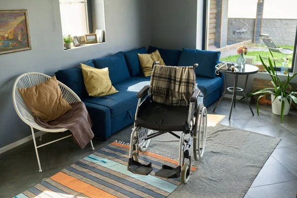 Wheelchair with woolen plaid standing on handmade rug in the center of living room surrounded by comfortable couch and armchair with cushions