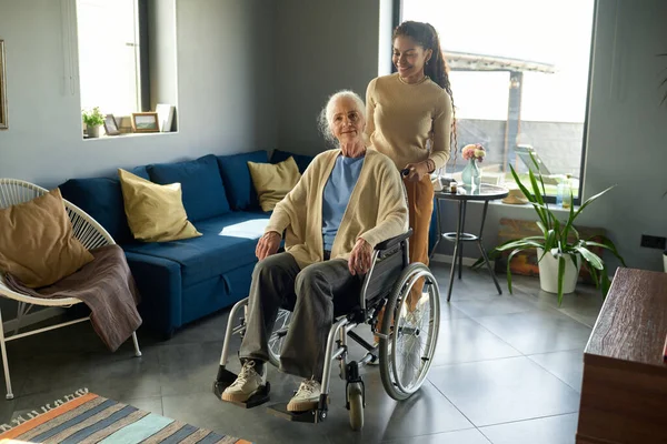 少女和祖母一起推着轮椅行走在宽敞的客厅里 有舒适的沙发和扶手椅 — 图库照片