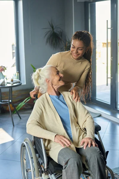 車椅子に座っている高齢女性ながら 障害のある幸せな祖母と孫娘がリビングルームでお互いを見て — ストック写真