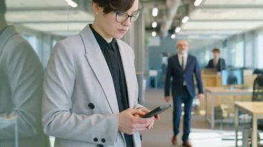 Resmi giyinmiş iş kadınının ofiste dikilirken ve iş günü akıllı telefondan mesaj yazarken görüntüsünü kaldır
