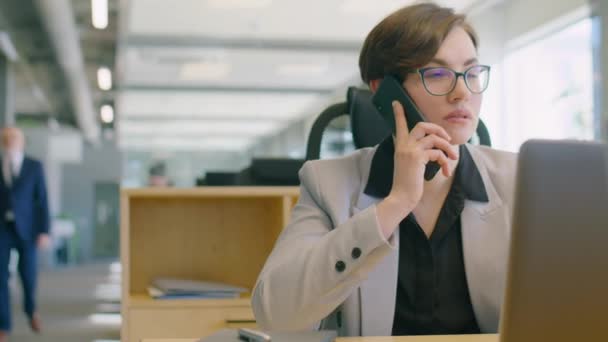在办公室的办公桌前工作时 穿着正装 戴眼镜 用笔记本电脑和讲手机的女商人 — 图库视频影像