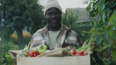 Orta boy Afro-Amerikan bir adamın elinde taze sebzelerle kasayla bahçede dikilirken gülümsediği bir fotoğraf.