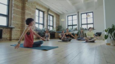 Yoga dersi sırasında kadın eğitmenle birlikte spor giyim kuşağı giymiş, minderlerde omurga bükme çalışması yapan bir grup kadının fotoğrafını yakınlaştır.