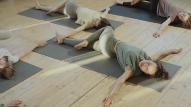 Spor kıyafetli bir kadının yüksek açılı pozu, sırt üstü uzanarak esneme egzersizi yapması ve grup yogası sırasında sırtüstü uzanan çocuk pozu alması.