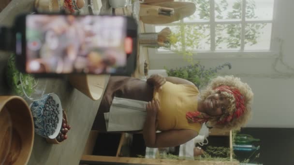 キッチンで三脚にスマートフォンでフードブログを撮影しながら カメラで話してエプロンでアフリカ系アメリカ人女性の垂直選択的フォーカスショット — ストック動画