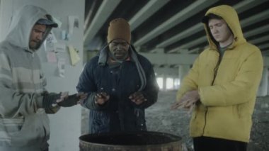 Bir grup çoklu etnik kökenli evsiz adam, terk edilmiş binada yan yana dururken, yanan ateşin üzerinde ellerini ısıtıyorlar.
