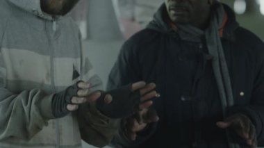 Afrika kökenli Amerikalı ve Kafkasyalı evsiz adamların ateş fıçısının üstünde ellerini ısıtırken ve konuşurken çekilmiş el kamerası görüntüleri.