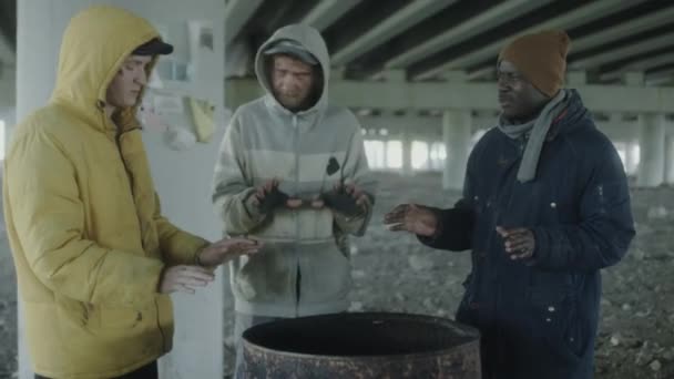 Три Багатоетнічні Безпритульні Чоловіки Розігрівають Руки Над Вогневим Стволом Обговорюють — стокове відео