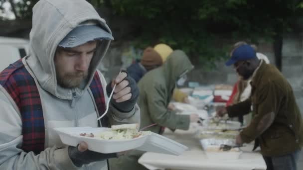 一个留着胡子的无家可归的人在城市街道上的厨房旁边吃着免费的塑料容器食物 — 图库视频影像