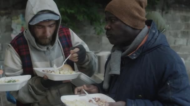 屋外スープキッチンでボランティアが提供する無料の食事を食べながら話している多民族ホームレスの男性のグループ — ストック動画