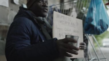 Afro-Amerikalı evsiz gazi elinde karton tabela ve metal kutuyla sokakta oturuyor ve insanlar bağış yaparken para dileniyor.