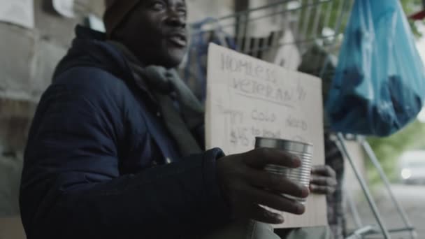 アフリカ系アメリカ人のホームレスのベテランが段ボールの看板と金属缶を持って路上に座って寄付をする人々としてお金を請う — ストック動画