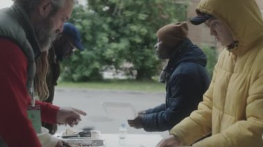 Çok ırklı evsiz insanlar gönüllülerden bedava yemek alıyor ve konteynırlardan yemek yiyorlar.