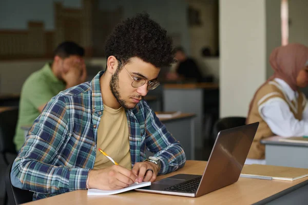 ノートパソコンを持って机の上に座ってノートを作る若者 彼はクラスで言語を学ぶ — ストック写真