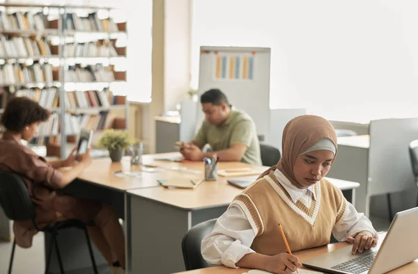 年轻的穆斯林女孩在与其他有背景的移民一起坐在图书馆时 用笔记本电脑学习外语 — 图库照片