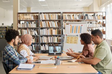 Bir grup yabancı öğrenci kütüphanede otururken öğretmenleriyle online ders alıyorlar.