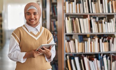 Kütüphanede tablet bilgisayar kullanırken kameraya gülümseyen Müslüman öğrencinin portresi