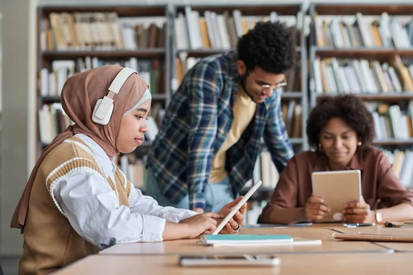穆斯林学生在图书馆与其他学生一起学习时 用无线耳机听平板电脑上的在线课程 — 图库照片