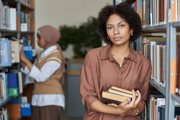 Portrait of African AMerican student looking at camera while standing with books near the bookcase in library
